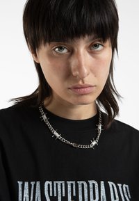 Collana in argento con punte e dettagli a spirale, indossata sopra una maglietta nera con un audace testo grafico bianco.