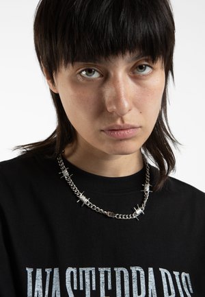 Zilveren ketting met spikes en opgerolde accenten, gedragen over een zwart T-shirt met gedurfde witte grafische tekst.