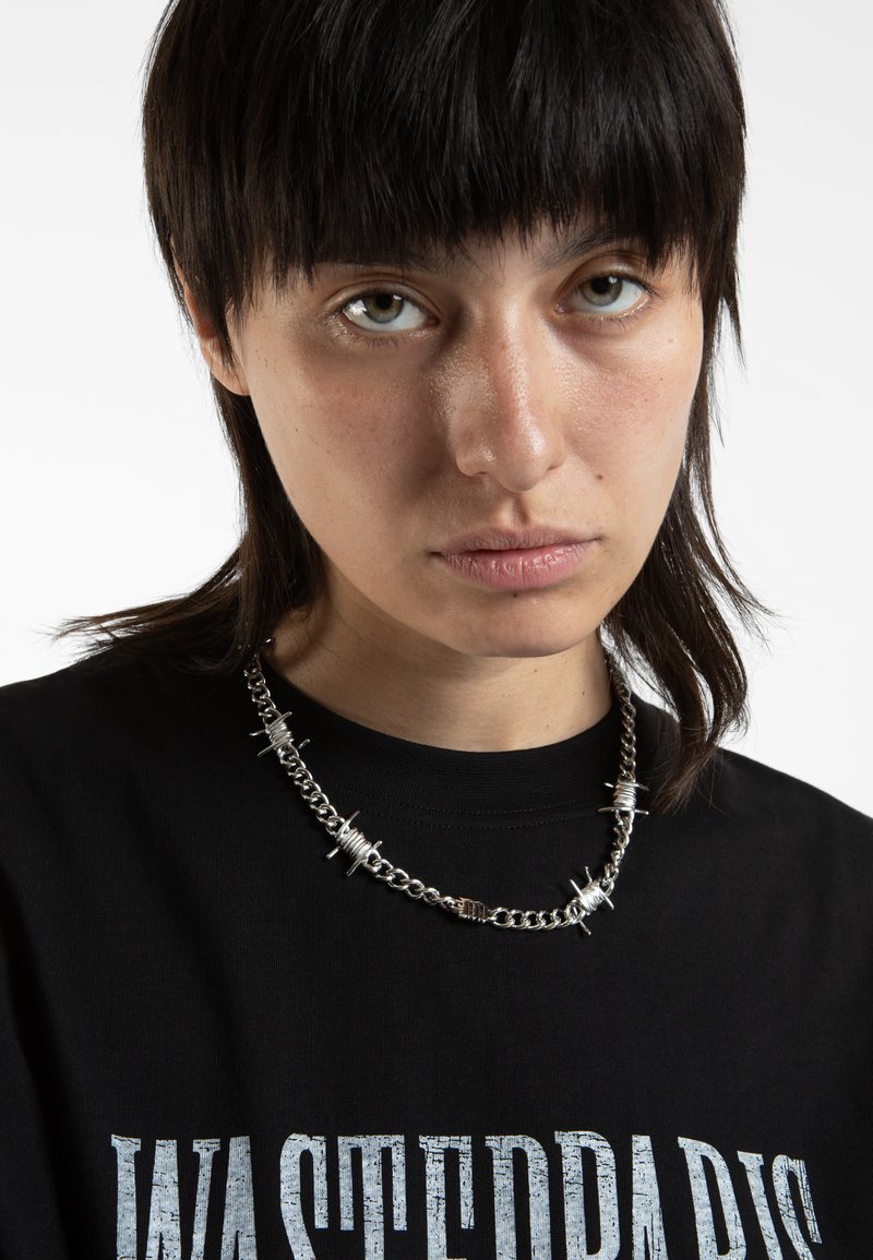 Collana in argento con punte e dettagli a spirale, indossata sopra una maglietta nera con un audace testo grafico bianco.