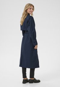 Marineblauwe lange jas met een cropped ontwerp, ceintuur in de taille en zijsplitten. Gemaakt van gladde stof, met lange mouwen en een klassieke kraag.
