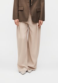 Pantalon beige à jambes larges avec plis, associé à un blazer marron cintré. Les chaussures présentent des talons pointus gris clair.