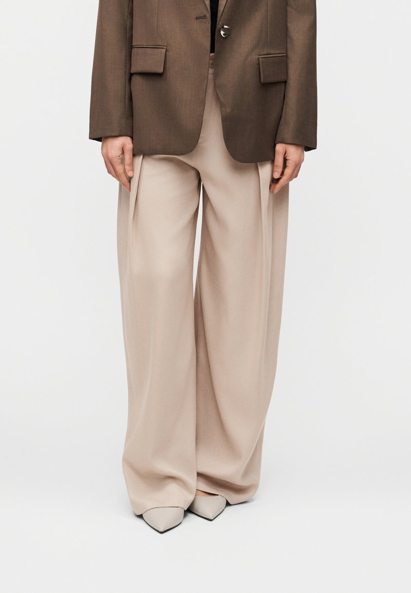 Pantalon beige à jambes larges avec plis, associé à un blazer marron cintré. Les chaussures présentent des talons pointus gris clair.