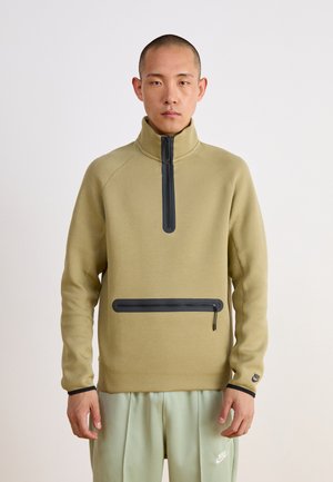 Hombre vestido con sudadera beige de cremallera con dos cremalleras negras y pantalones Nike verdes claros, de pie frente a un fondo blanco liso.