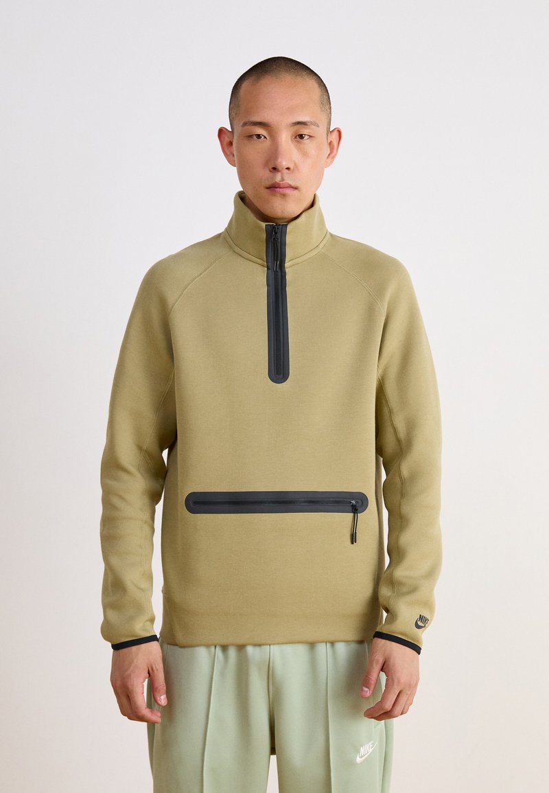 Άνδρας με μπεζ φούτερ με φερμουάρ και δύο μαύρα φερμουάρ, φορώντας ανοιχτό πράσινα παντελόνια Nike, στέκεται μπροστά σε λευκό φόντο.