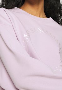 Lavendel sweatshirt med subtilt glansigt cirkulärt mönster över bröstet, buren av en person med mörkt, vågigt hår och en silverhalsband.