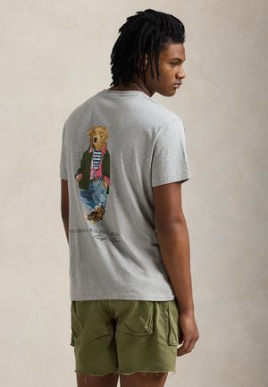 Fiatal férfi rasztahajjal, szürke pólót visel, amin egy réteges öltözékű medve látható, és olívazöld cargo rövidnadrágot hord.