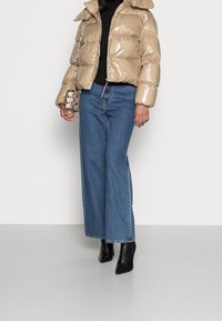 Beżowa błyszcząca kurtka puchowa z wysokim kołnierzem, w zestawieniu z szerokimi niebieskimi dżinsami i czarnymi spiczastymi botkami za kostkę. W ręku mała torebka w wzorki.