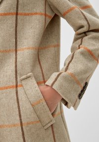 Abrigo de lana beige con un patrón de espiga, con cuadros en naranja y marrón, un bolsillo lateral y detalles de botones en el puño.