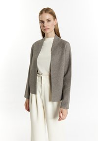 Cardigan gri deschis cu față deschisă și mâneci lungi, realizat dintr-un material moale. Asortat cu un pulover crem cu guler înalt și pantaloni albi cu talie înaltă.