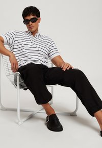 Pasiasta biała koszulka polo z kołnierzykiem, czarne spodnie ze ściągaczami, oraz czarne buty, siedząca na białym krzesle z drutu.