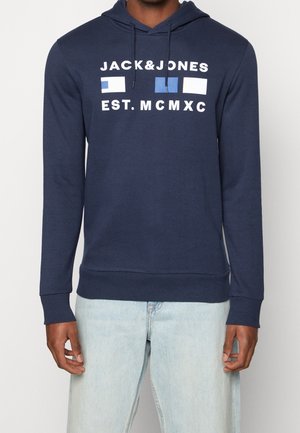 Mand iført navyblå hættetrøje med "JACK&JONES EST. MCMXC" og tre rektangulære farveblokke, kombineret med lyseblå jeans.