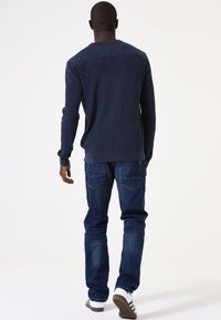 Navyfarbener, strukturierten Pullover mit einem gerippten Muster und einem glatten Schulterdetail, kombiniert mit blauen Jeans und weiß gestreiften Schuhen.
