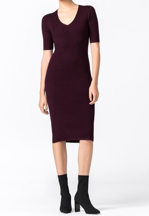 Frau trägt ein eng anliegendes, knielanges Pflaumenkleid mit kurzen Ärmeln und schwarzen, hochhackigen Stiefeletten, steht vor weißem Hintergrund.