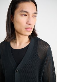 Svart nätcardigan med v-ringning, featuring knäppning och långa ärmar. Bärs över en figursydd svart ribbad tröja som visar upp intrikata texturer.