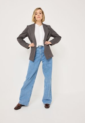 Blazer grigio in lana su una camicetta bianca con bottoni, abbinato a jeans in denim blu a vita alta e a gamba larga, e scarpe con tacco marroni scuri.