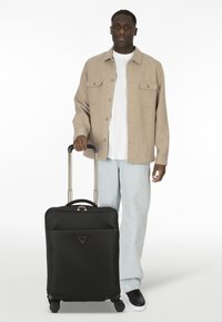 Schwarzer Rollkoffer mit strukturierter Oberfläche, Metallbeschlägen und einem Logo. Verfügt über einen ausziehbaren Griff und vier Rollen für Mobilität.