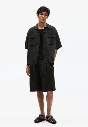 Schwarzes, strukturiertes Kurzarmhemd mit zwei Brusttaschen, kombiniert mit passenden knie-langen Shorts und schwarzen Riemchensandalen.