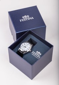 Montre argentée avec un cadran blanc, un bracelet en cuir noir et des accents argentés, présentée dans une boîte cadeau bleue avec un logo de marque.