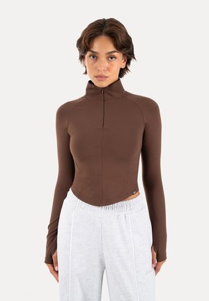 Mujer con el pelo corto y castaño que lleva una camiseta ajustada marrón de manga larga con cremallera frontal y pantalones blancos con cordón, de pie frente a un fondo liso.