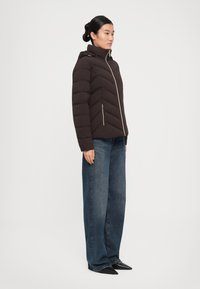 Kvinne som står iført en mørk brun quiltet jakke med skråsting, blå vide jeans og svarte spisse sko.