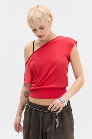 Žena s krátkymi blond vlasmi nosiaca červený top bez ramienok, čierne nohavice s reťazovým opaskom a strieborné náramky, pozerajúca dole.