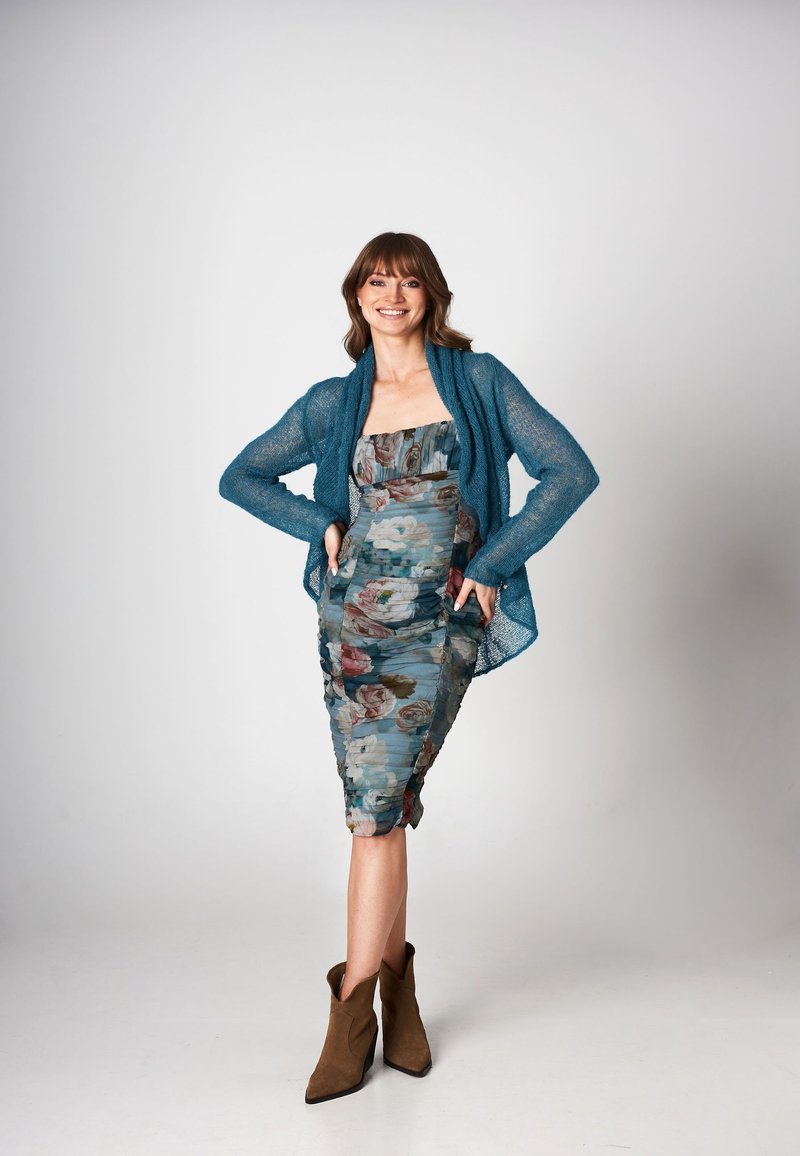 Szmaragdowy sweter w splot, sukienka w kwiaty w odcieniach niebieskiego i różowego, dopasowany krój, długość do połowy łydki oraz brązowe botki za kostkę.