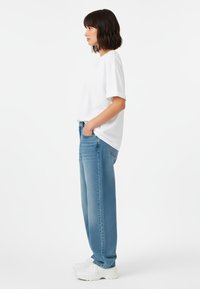 Camicia bianca a maniche corte abbinata a jeans azzurri a vestibilità ampia e sneakers bianche, in piedi su uno sfondo neutro.