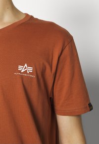 Kurzarm-T-Shirt aus verbranntem Orange aus Baumwolle. Mit weißem Logo und Text auf der Brust sowie einem schwarzen Etikett am Ärmel.