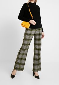 Svart texturerad topp, rutiga grå byxor med gula linjer och en liten handväska med krokodilprägling i gult. Svarta klackskor fulländar outfiten.