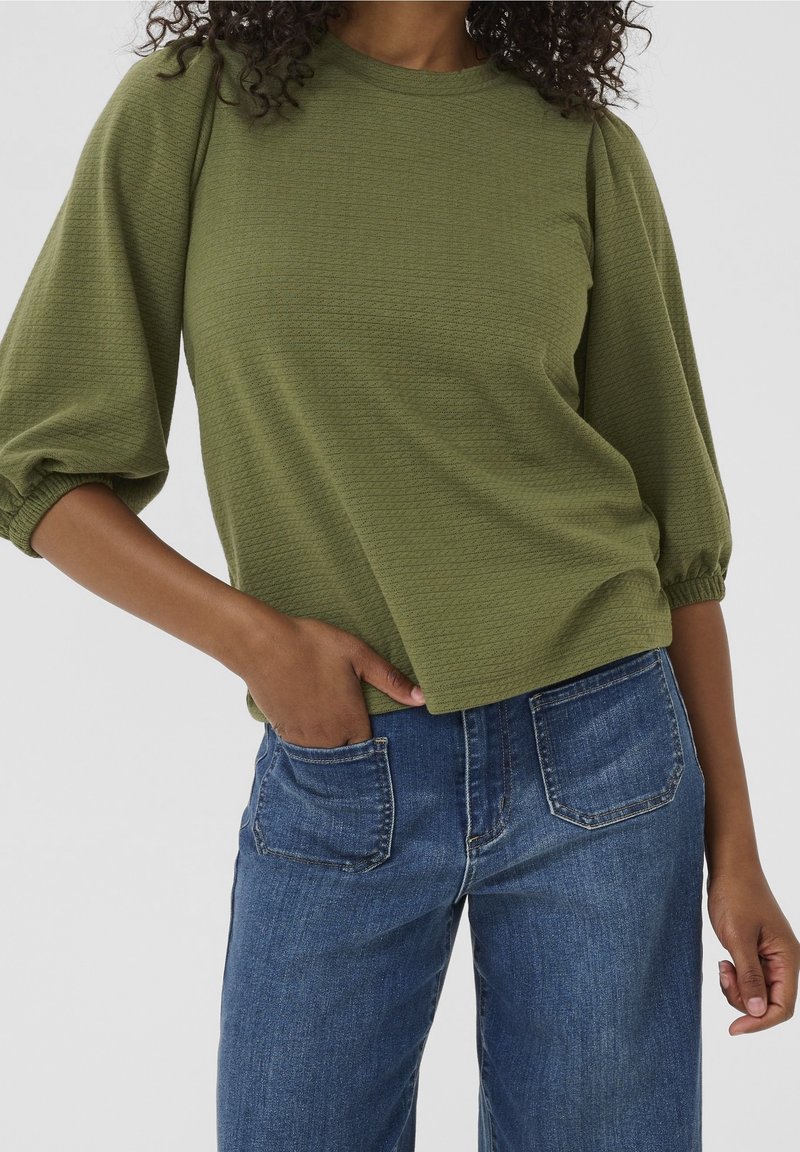 Femme portant un haut texturé vert olive à manches 3/4 et un jean en denim bleu, main dans la poche avant, sur fond uni.