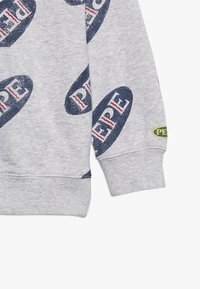 Szary rękaw i kieszeń bluzy z kapturem z powtarzającymi się napisami "PEPE" w granatowym i czerwonym kolorze oraz małą owalną łatką "PEPE" w pobliżu mankietu.