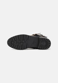 Suela de bota negra con un patrón de tracción robusto y una marca en relieve, mostrando un acabado texturizado con acentos de hardware en tono dorado.