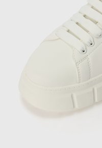 Weißer Sneaker aus glattem Synthetikmaterial, mit runder Zehenpartie, flachen weißen Schnürsenkeln und einer strukturierten Gummisohle mit dezenten Rillen.