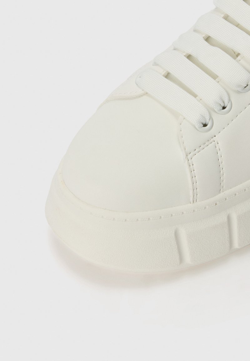 Weißer Sneaker aus glattem Synthetikmaterial, mit runder Zehenpartie, flachen weißen Schnürsenkeln und einer strukturierten Gummisohle mit dezenten Rillen.