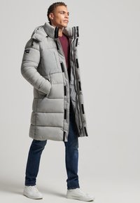 Abrigo acolchado gris claro con cuello alto y detalles en negro, con un patrón acolchado. Se lleva sobre una camisa granate y combinado con vaqueros.