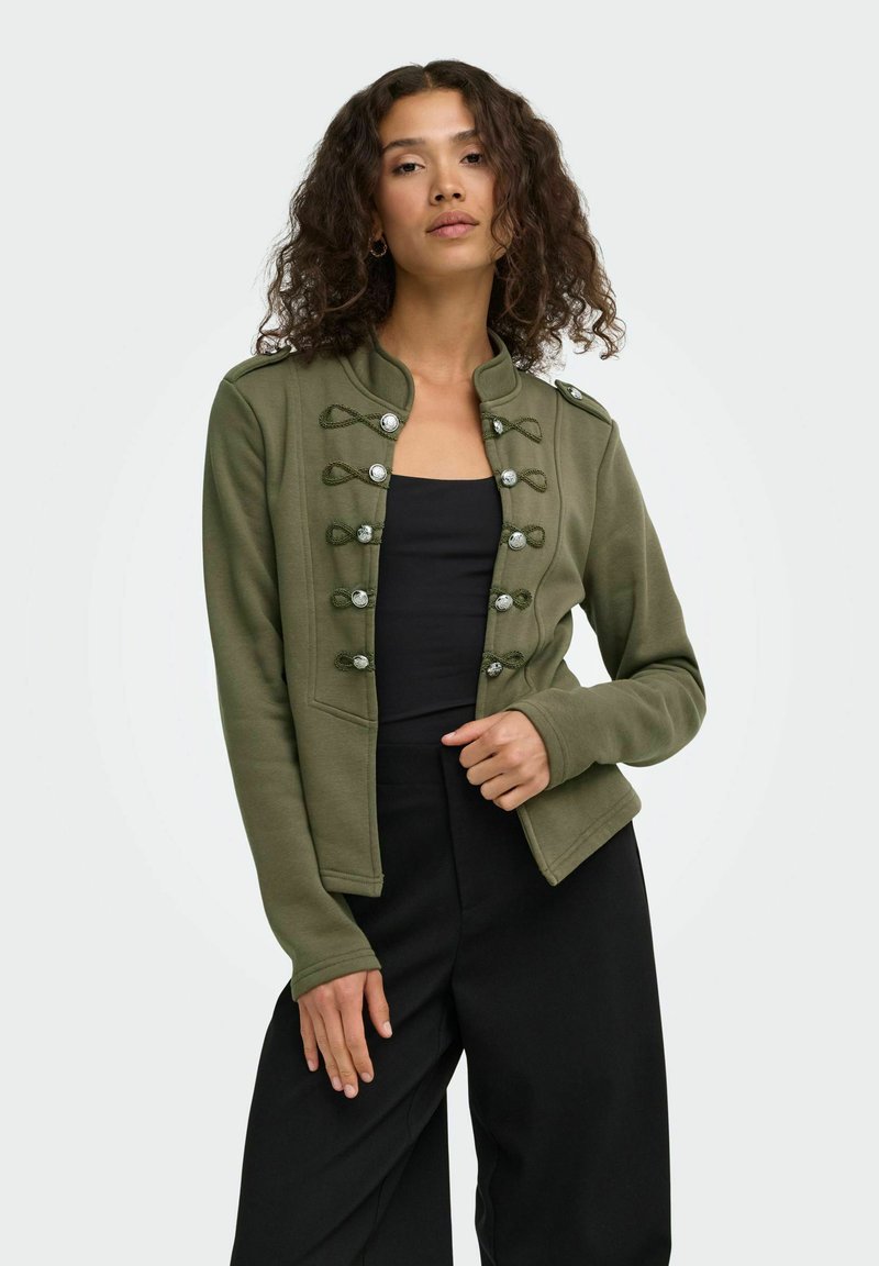 Femme aux cheveux bouclés portant une veste militaire vert olive sur un haut noir et un pantalon noir, debout devant un fond uni.