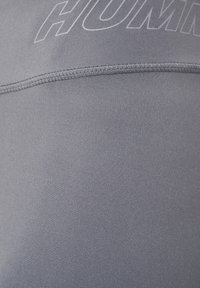 Débardeur de sport gris avec une texture lisse, des coutures à plat et un logo bien en évidence sur la partie supérieure. Le matériau semble extensible.