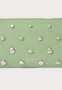 Gaiši zaļa ādas somiņa ar sudraba krāsas dome formas kniedēm vienmērīgi izvietotām, rāvējslēdzēja aizdare augšpusē un nospiestu "kate spade NEW YORK" logotipu apakšā.