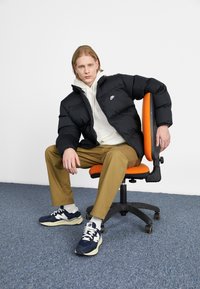 Sort pufferjakke, hvid hættetrøje under, khaki bukser og marineblå sneakers. Sidder på en orange kontorstol med en grå tæppebaggrund.