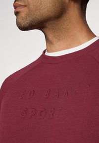 Felpa bordeaux con lettering testurizzato "Ted Baker Sport", scollo a giro, colletto interno bianco a contrasto e maniche raglan.