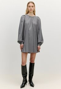 Graues langärmliges Kleid mit Rundhalsausschnitt und gerafften Schultern, kombiniert mit knielangen schwarzen Stiefeln mit spitzem Zehenbereich. Glatte Stofftextur.