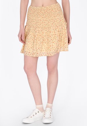 Frau trägt einen kurzen, hellgelben Rock mit Blumenmuster und geraffter Taille, durchsichtige Knöchelsocken und weiße Schnürsneaker, steht vor weißem Hintergrund.