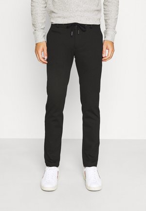 Hombre con pantalones negros ajustados con cintura de cordón, camiseta gris de manga larga y zapatillas blancas, de pie sobre un suelo claro y liso.