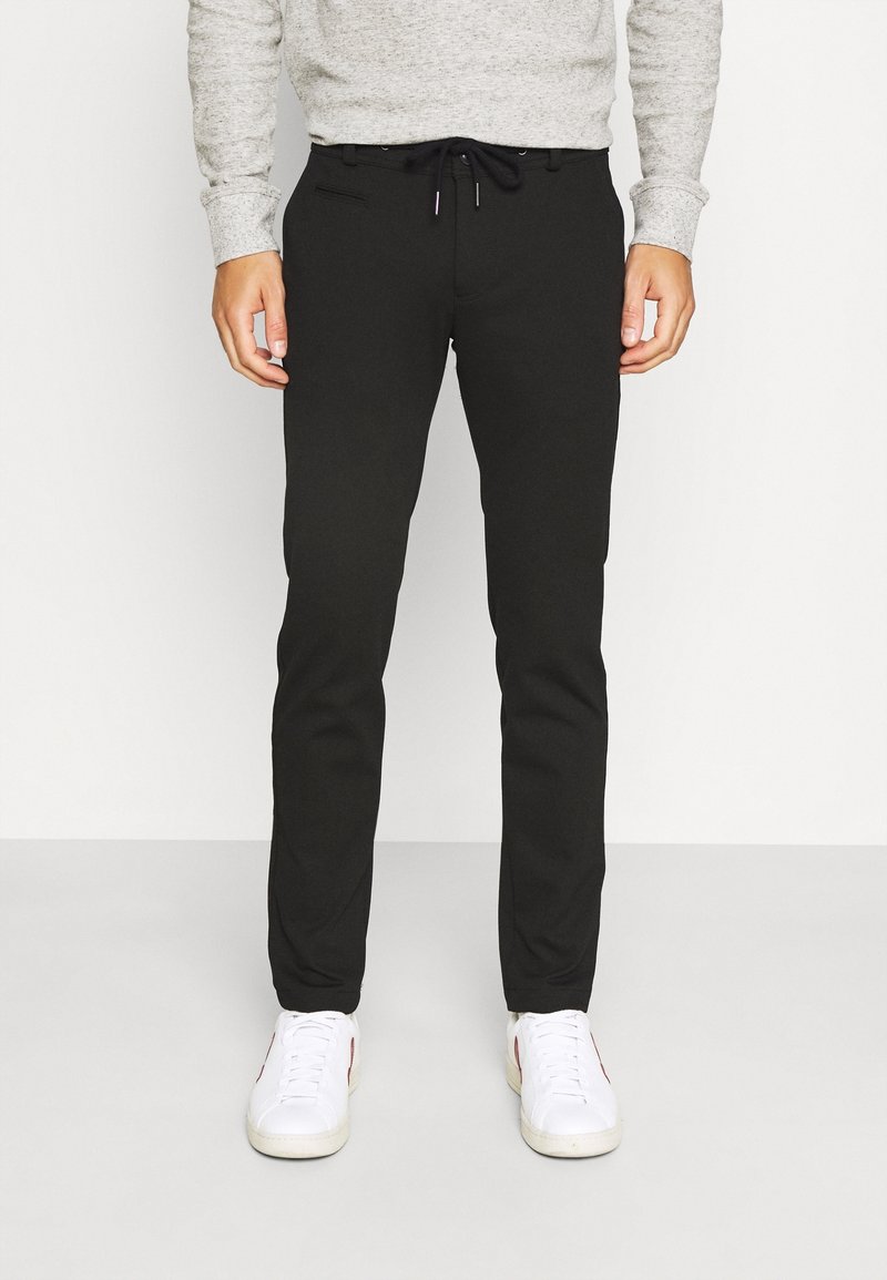 Homme portant un pantalon noir slim à taille à cordon, une chemise grise à manches longues et des baskets blanches, se tenant sur un sol clair uni.