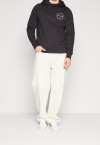 Uomo che indossa una felpa nera Tommy Hilfiger, pantaloni larghi color avorio e sneaker bianche Adidas con strisce nere, in piedi davanti a uno sfondo semplice.