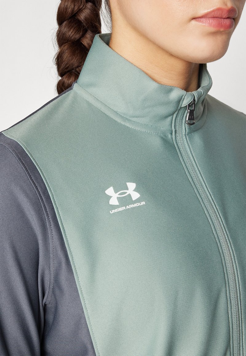 Giacca sportiva verde chiaro con zip e colletto alto, maniche grigie. Presenta il logo Under Armour bianco sul petto. Tessuto testurizzato.