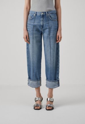 Jeans de denim azul claro con piernas anchas, dobladillos enrollados y costuras en contraste. Combinados con sandalias metálicas con grandes hebillas.