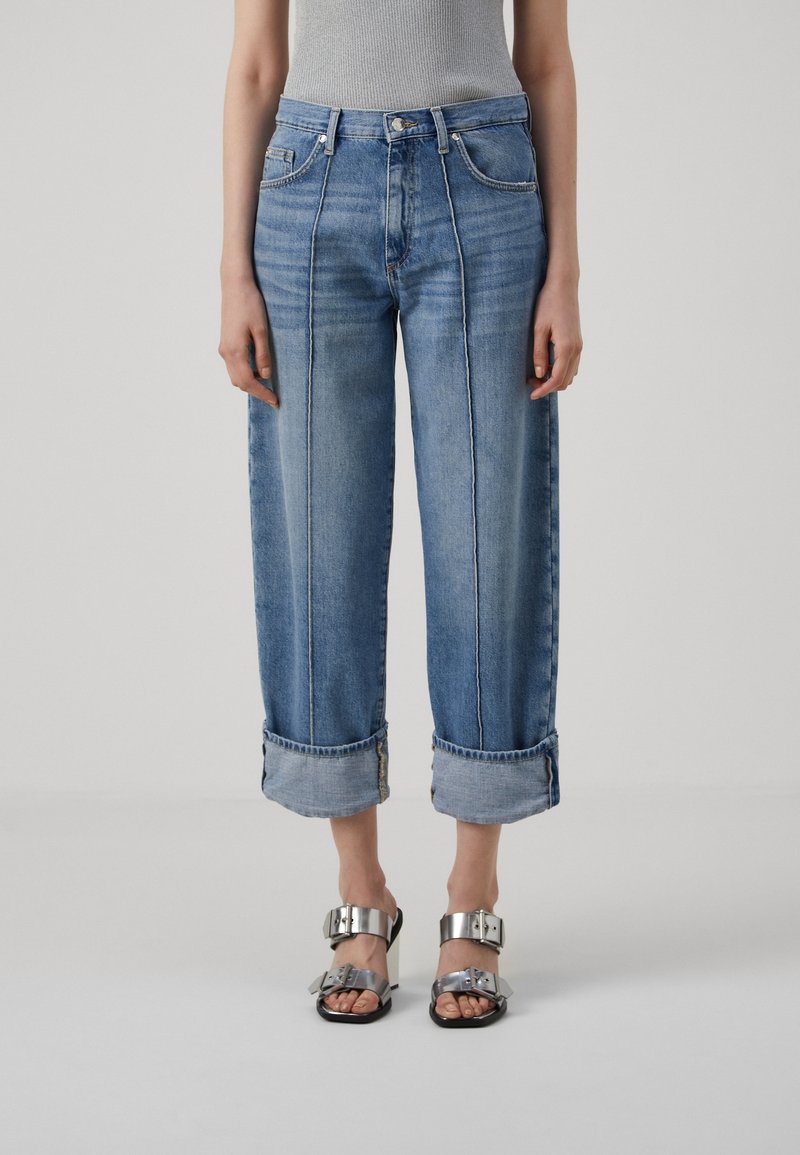 Jeans de denim azul claro con piernas anchas, dobladillos enrollados y costuras en contraste. Combinados con sandalias metálicas con grandes hebillas.