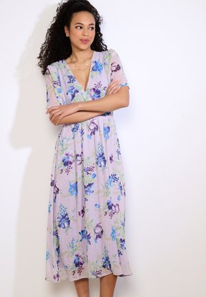 Žena s kučeravými vlasmi v levanduľových kvetovaných midi šatách s krátkymi rukávmi, stojaca so založenými rukami na bielom pozadí.