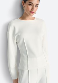 Valge pikkade varrukatega särk ümmarguse kaelusega ja struktureeritud õmblustega, millel on lõikevolditud siluett ja pehme tekstiilipind.